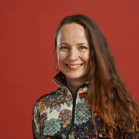 Ruskeahiuksinen nainen hymyilee kameralle hiukset auki. Päällään värikäs paita ja korvakorut. Kuvassa Minna Nerg.